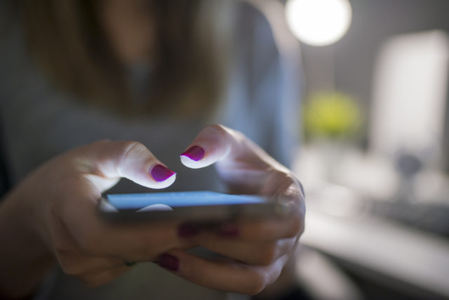 Front view of a woman pointing finger to smartphone with white monitor in the hands, working a computer keyboard on table. Business, technology and people concept - close up of Unrecognizable businesswoman with black mobile phone while sitting gin home office at night.