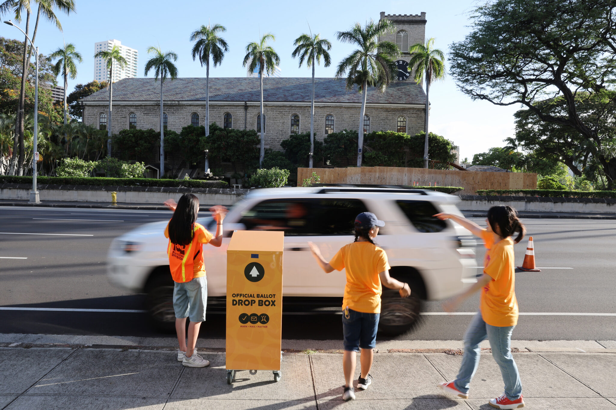 Roosevelt HS students Sophie Lo, 16, (L-R) Emily Li, 17, and Ying Cai, 17, wave to drivers as they collect their ballots at Honolulu Hale, Saturday, August 10, 2024 in Kapolei, Hawaii. Marco Garcia/CivilBeat/2024