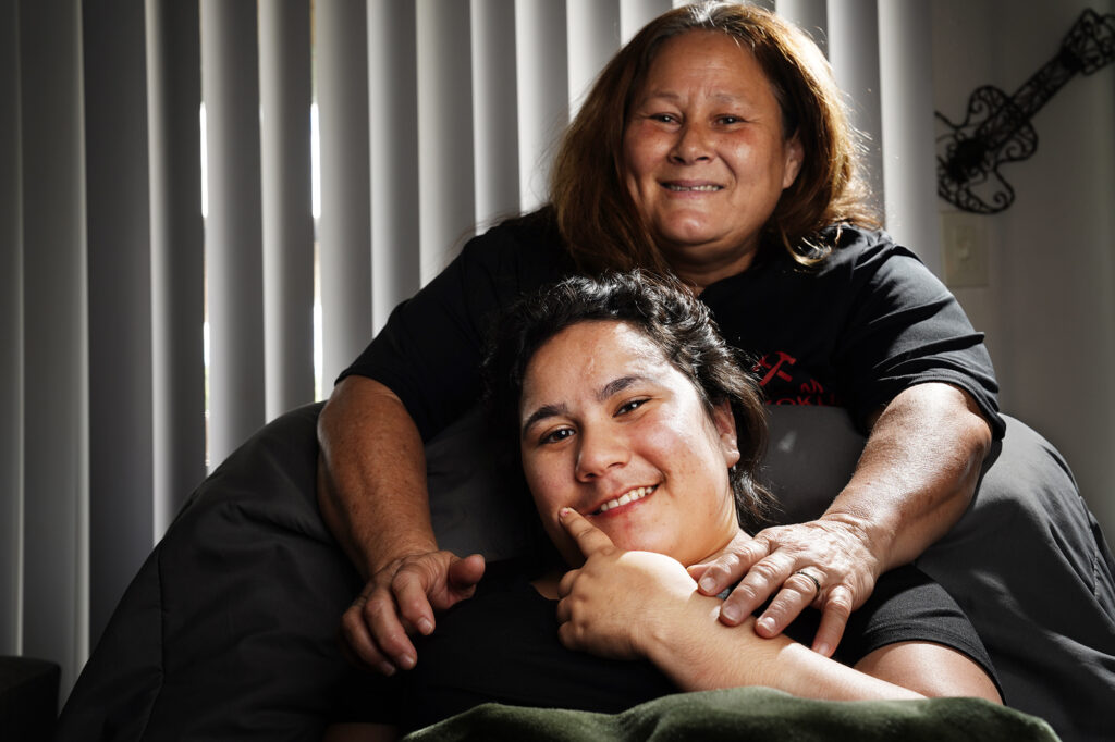 Richianna DeGuzman, 18, and her mother Susan Mahiai pose for a portrait Friday, Oct. 4, 2024, in Ewa Beach. DeGuzman was shot in the leg and head on June 17, 2023, in Maiili. She currently must lift the right side of her face to smile. The bullet damaged the left side of her brain. Her mother says she was caught in a gunfire exchange between gangs. (Kevin Fujii/Civil Beat/2024)