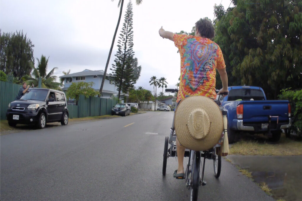 A neighbor flashes a shaka to woodturner Aaron Hammer on his custom-outfitted tricycle Wednesday, July 2, 2025, in Kailua. The trike is equipped with woodturning lathe, storage for tools, supplies and battery making it a mobile art studio. Still image taken from video. (Kevin Fujii/Civil Beat/2025)