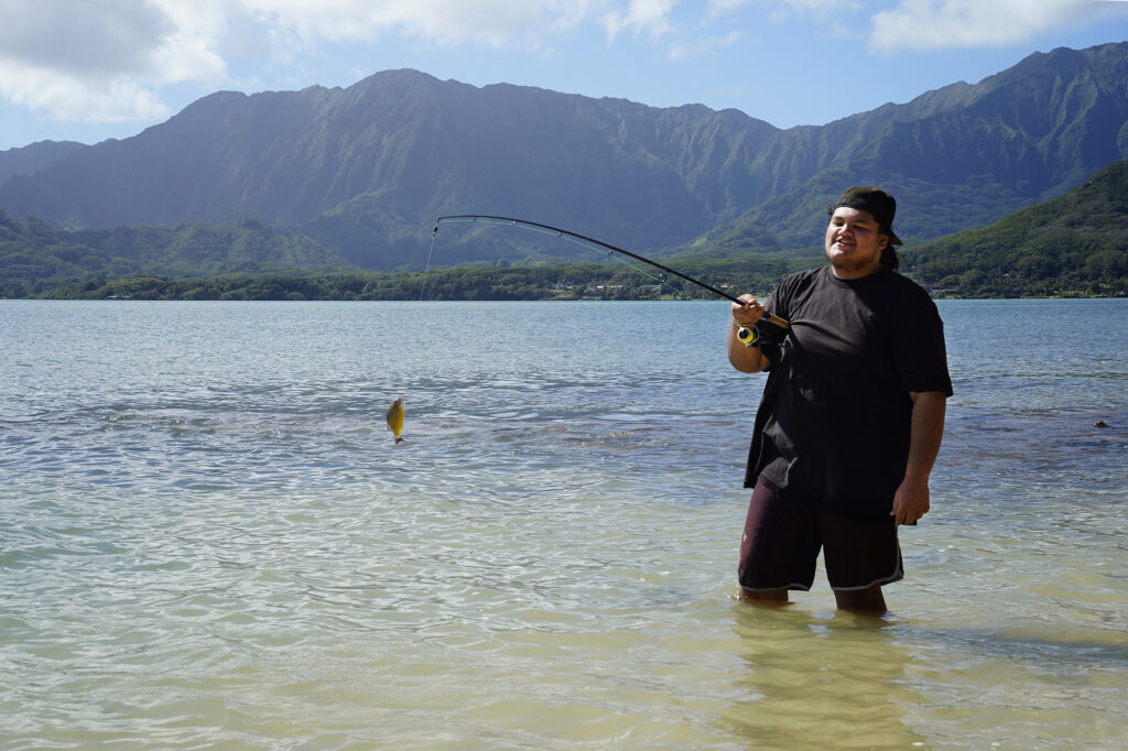 Hakipuu Charter School senior Aka Kahuhu laughs after reeling in a small fish believed to be an invasive Ta'ape, or common bluestripe snapper, identical to the one he just threw back in the water Monday, Nov. 24, 2025, at Kualoa Regional Park in Kāneʻohe. Fishing is part of Hinkel’s science class. (Kevin Fujii/Civil Beat/2025)