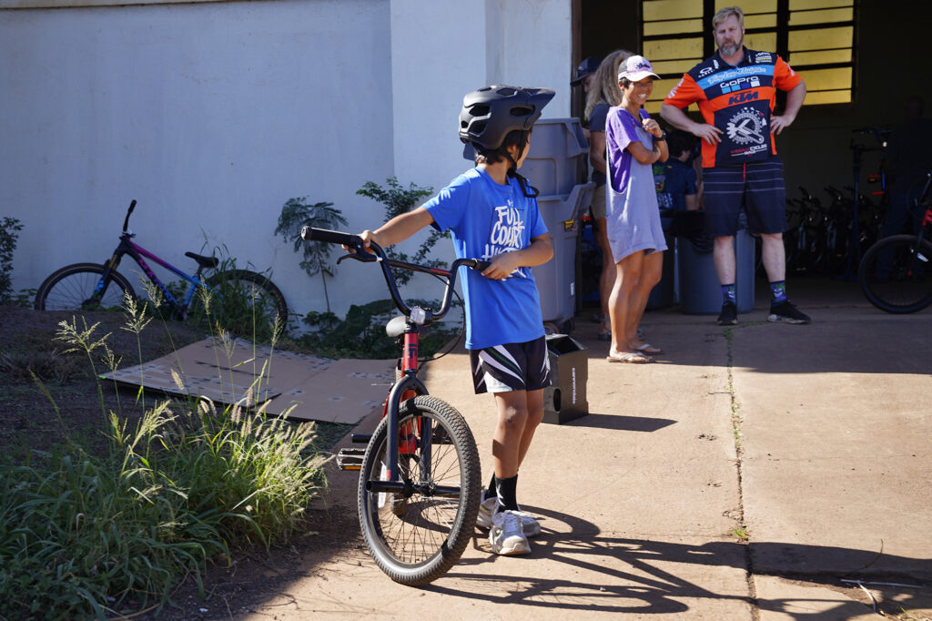 Elisha Oshima, 9, looks back toward his mother Mayumi and Krank Cycles Maui owner Aaron ”Moose" Reichert during Jim Falk’s annual Christmas Kids Bike Build Saturday, Dec. 6, 2025, in Puʻunēnē. Approximately 450 new bicycles are built and distributed in Maui County. Oshima and his family have been volunteering with the bike build and their church. (Kevin Fujii/Civil Beat/2025)