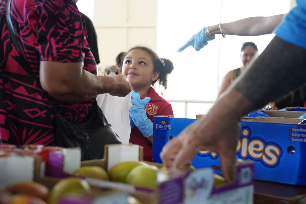 Kuhio Public Housing food pantry volunteer Elliana McMore, 5, offers tangerines to a resident Tuesday, Dec. 9, 2025, in Honolulu. (Kevin Fujii/Civil Beat/2025)