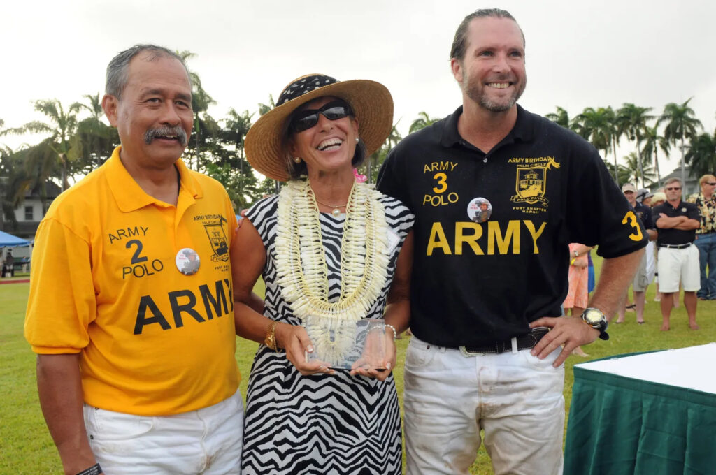 A woman and two men stand outdoors, smiling at the camera. The woman holds a trophy, and the men both wear shirts that say “Army” and “Polo.”