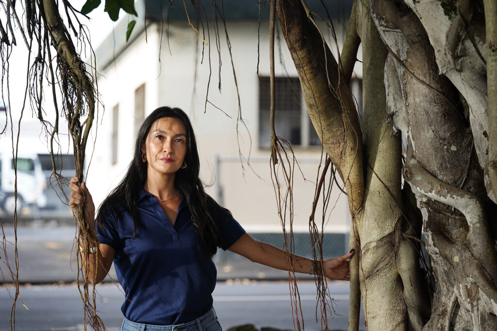 Ashley Kierkiewicz is photographed under a large banyan tree at Kalākaua Park Tuesday, Dec. 16, 2025, in Hilo. She grew up spending much of her childhood in the building behind her. It was her father’s Hilo Auto and Truck Service station. She bemoans its derelict state and fencing because of the homeless in the area. (Kevin Fujii/Civil Beat/2025)