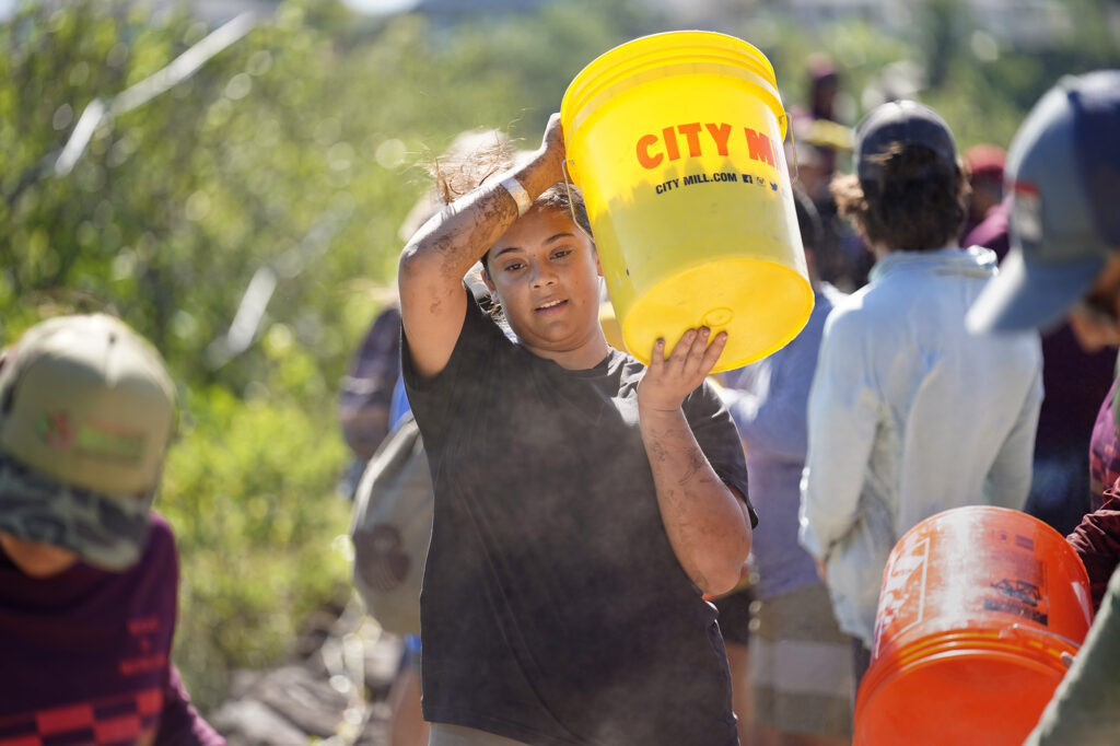 Moʻokuapā volunteer Isabelle Colorado carries a bucket of ko‘a (coral) pieces Saturday, Dec. 13, 2025, in Kāneʻohe. Colorado, who is stationed on Oʻahu with the military and came on her own, is one of 2,000 volunteers who joined the Moʻokuapā community work day to finish the wall of the 800-year-old He‘eia loko i‘a (fishpond). (Kevin Fujii/Civil Beat/2025)