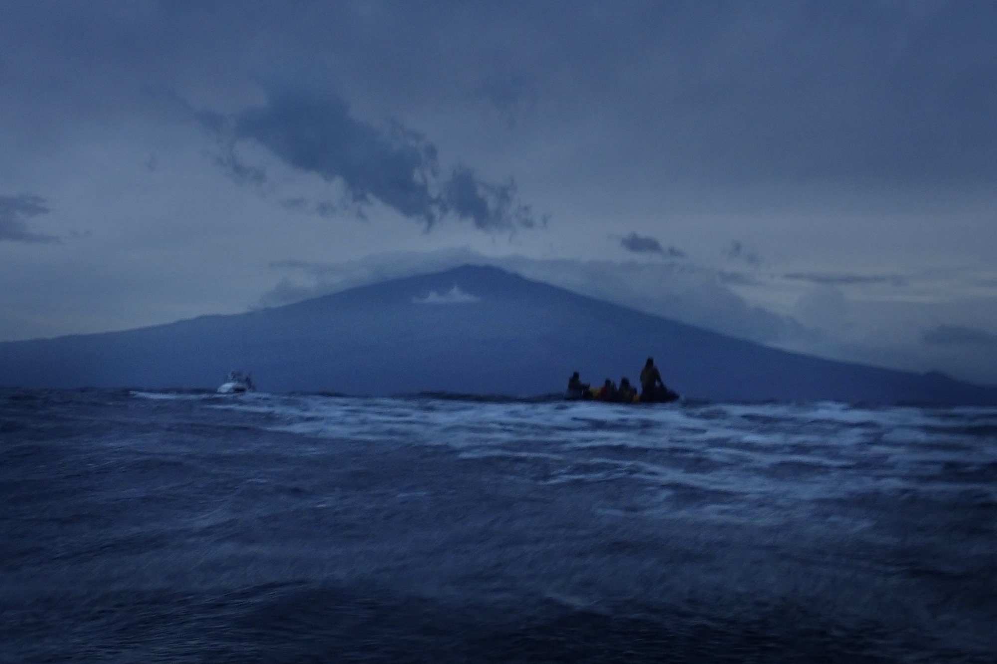 Protect Kahoʻolawe ʻOhana (PKO) lands at Hakioawa the the pre-dawn hour Monday, Jan. 5, 2026. This morning, the winds screamed through the ʻAlalākeiki Channel between 30-40 knots with wind-generated swells 10-12 feet. The 33-foot Pualele fishing boat to the left and the zodiac loaded with people and ukana (duffel bags, gear, coolers and buckets), right, times the waves before heading toward the line of helpers treading water or in shallower water. The group sailed from Māʻalaea Harbor and Kīhei Small Boat Harbor on the Pualele and Pualele Iki fishing boats. As the two larger boats neared Kahoʻolawe those aboard performed an oli (chant) requesting permission to step onto Kahoʻolawe. A smaller inflatable zodiac then pulled up alongside the larger fishing boats and responded with a welcoming oli. Then people and ukana transferred over in the rough seas. The zodiac motored as close as safely possible then people jumped into the water and handed ukana down a line. (Kevin Fujii/Civil Beat/2026)