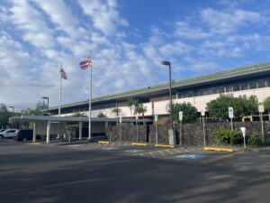 The front of the Hawaiʻi County building in Hilo taken from the parking lot. The Hawaiian and U.S flag wave at the entrance.