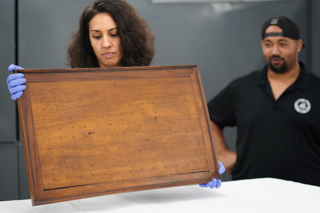 Kayla Kuʻualoha Fernandez, from left, Bishop Museum’s Ethnology Collections Manager, turns over the kūpuna papamu for Wallace Akua to check the base and feet Friday, Jan. 30, 2026, in Honolulu. This papamu is made of koa wood. It was donated to Bishop Museum in 1923 by Princess Elizabeth Kahanu as a part of the Kalanianaʻole-Kapiʻolani Collection. (Kevin Fujii/Civil Beat/2026)