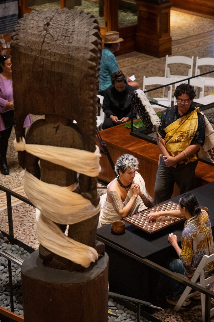 Kanaloa dressed for Makahiki watches over an all-Oʻahu championship match between Kapena Baptista, center, and Kahekili Yung, right, during the inaugural Ka Makou Kōnane Invitational Tournament in the center of the Bishop Museum’s Hawaiʻian Hall Friday, Feb. 13, 2026, in Honolulu. (Kevin Fujii/Civil Beat/2026)
