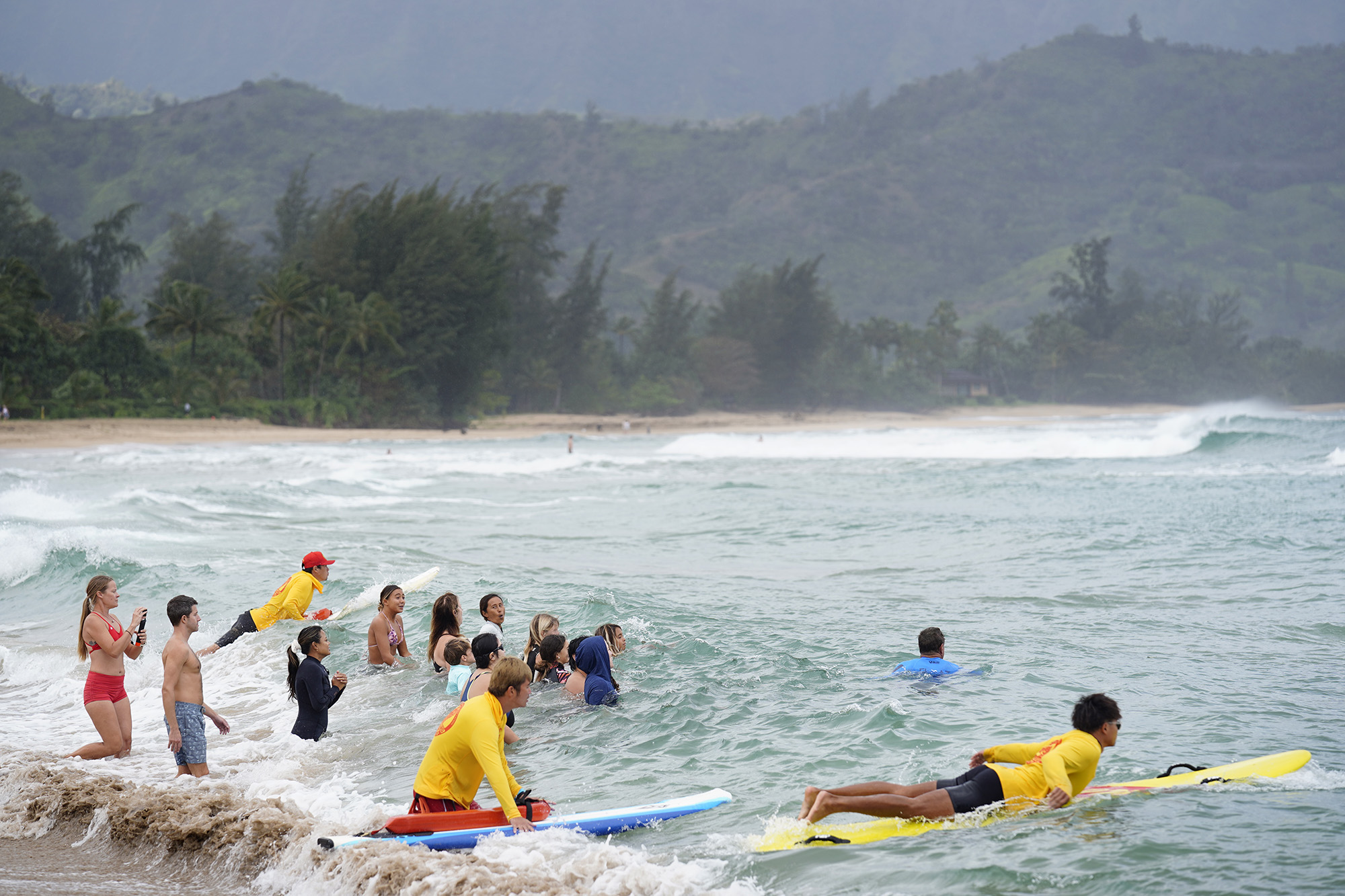 Ocean safety educator Bruce “Hoppo” Hopkins of Australia, wearing blue rash guard, top right, leads the second group of rip-current clinic participants purposefully into a Waiʻoli Beach Park rip current flanked by Okinawa Lifesaving Association lifeguards on rescue and surfboards Wednesday, Feb. 18, 2026, in Hanalei. (Kevin Fujii/Civil Beat/2026)