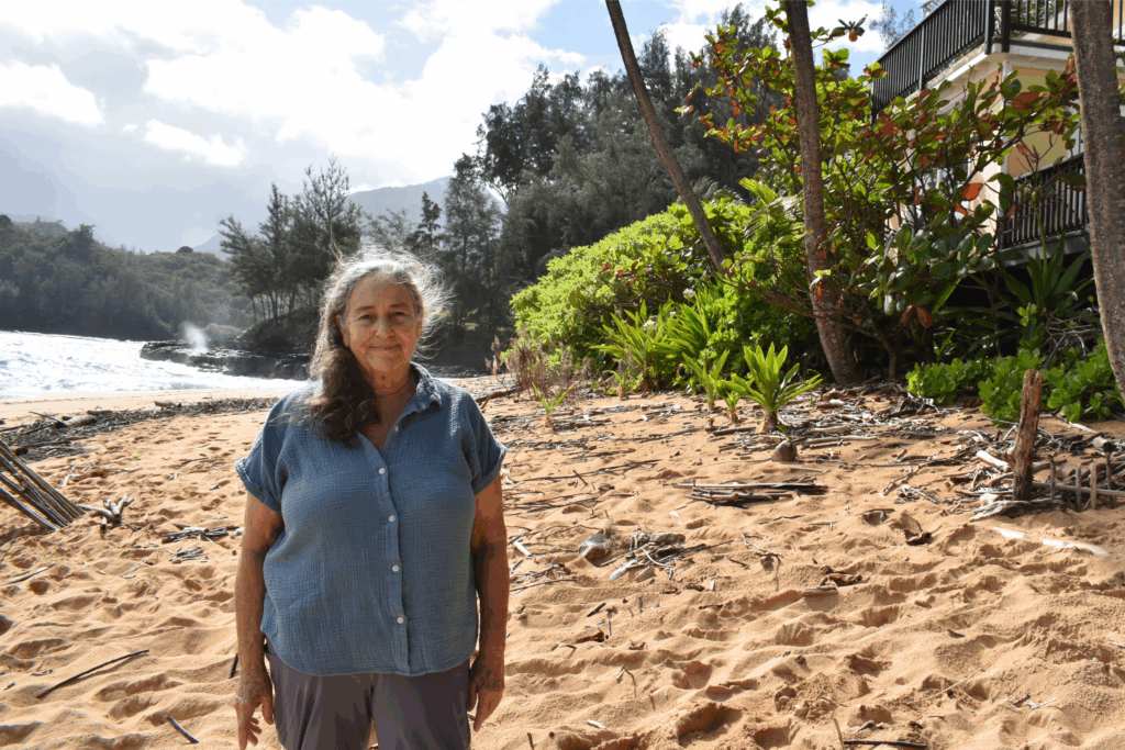 North Shore resident Caren Diamond stands in front of a pale yellow vacation rental at Wainiha Beach. Branches and other debris are scattered on top of the sand.