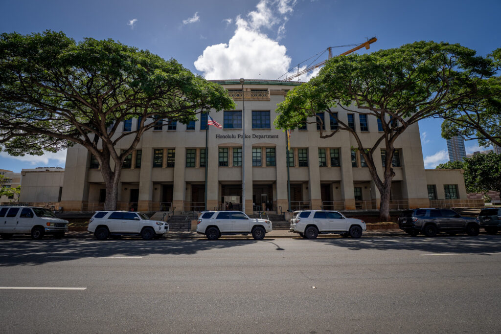 Honolulu Police Department headquarters building with a sign on the front of the building. Two large trees and several SUVs are parked in front of the building.