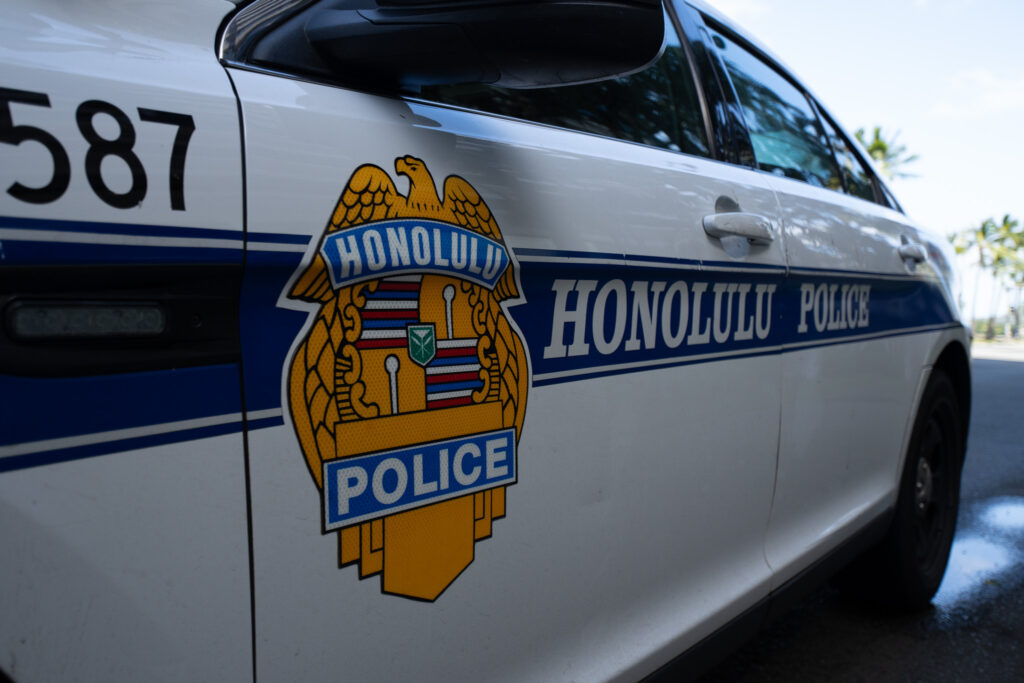 The side of a white Honolulu Police vehicle showing its golden orange logo and blue side stripes with “Honolulu Police” in white