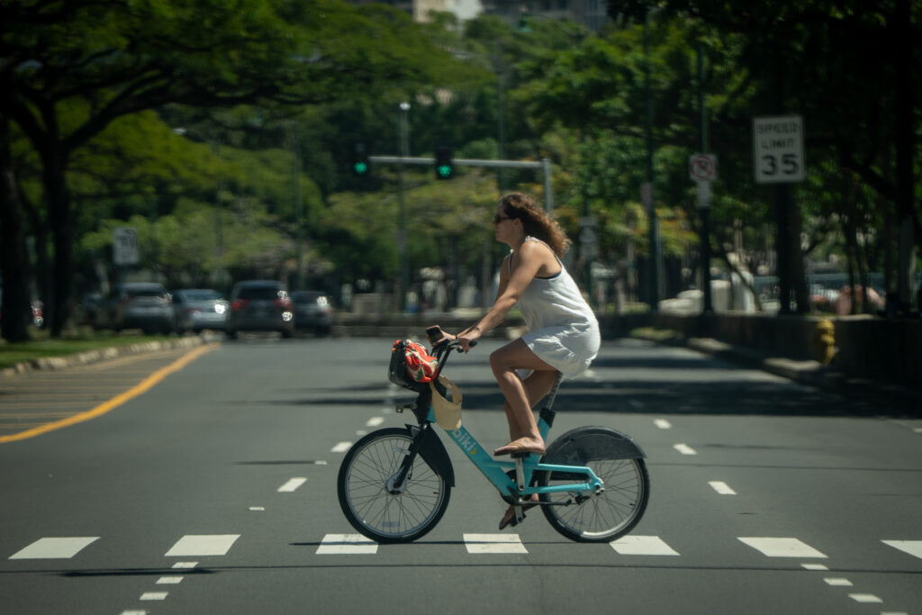A woman wearing a white outfit bicycles a blue Biki rental bike on a crosswalk. The roadway and foliage recede into the background.