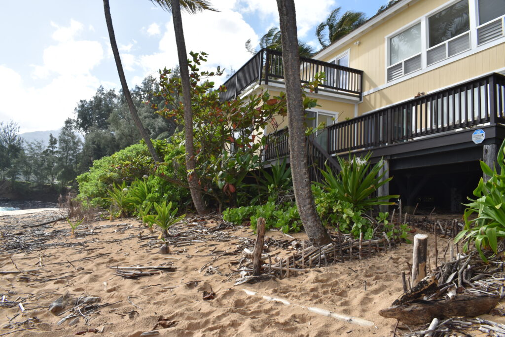 A pale yellow, two-story vacation rental house with outdoor lānai sits directly on Wainiha Beach. Branches and other debris lay scattered atop the sand.