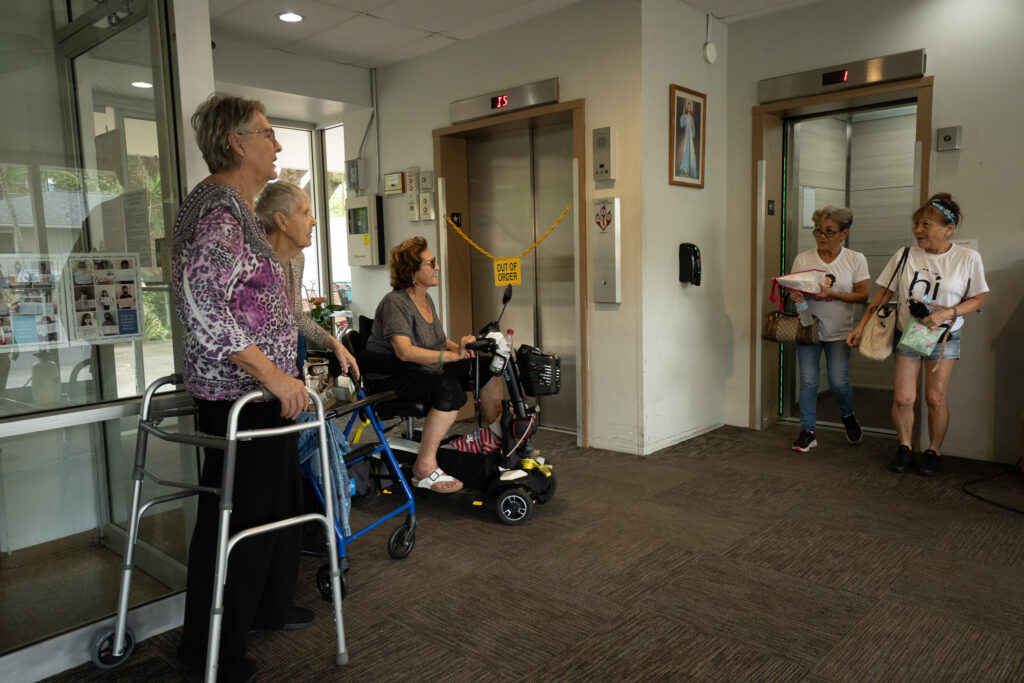Five women in the lobby of a residental apartment building waiting for elevator service. The women at left are using mobility devices such as walkers.