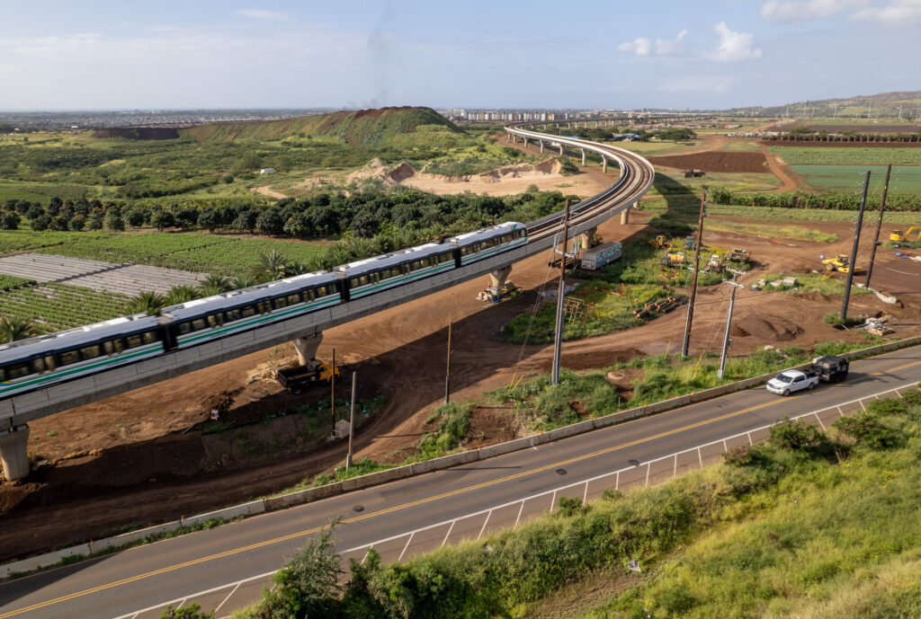 An elevated rail line and train are seen next to a roadway surrounded by farm and other less developed land.