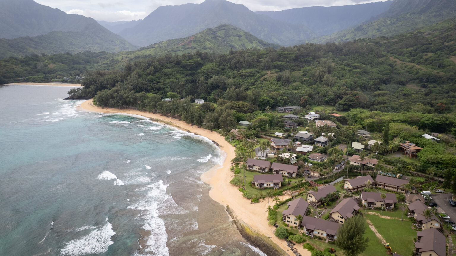 A light-colored strip of sand is seen in an aerial photograph that shows beachfront structures and mountainous land in the background.