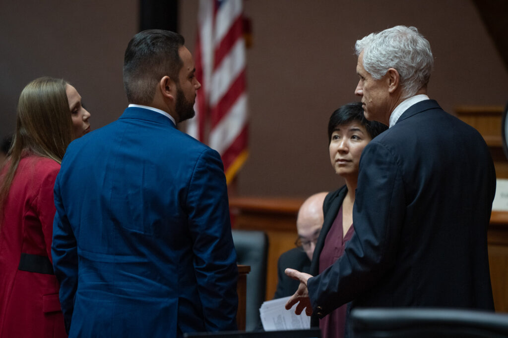 Four individuals speaking with one another: a woman in a red jacket with long brown hair; two men with blue jackets. The man on the left has dark hair and a close-cropped beard. The man on the right has white hair. The woman second from right has black hair. A balding man, seated is partially obscured.