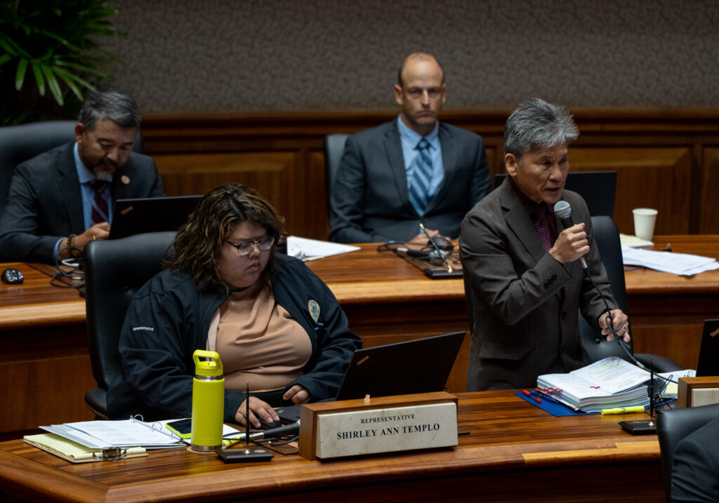 A man with a gray suit and graying hair holds a microphone, right, as other individuals sit around him. A woman sits to the left and two men are seated wearing jackets and ties in the background.