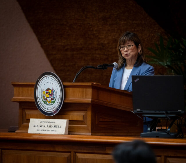 A woman with a light blue blazer, dark hair and glasses speaking at a lecturn