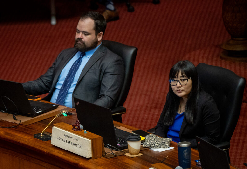A man and woman seated at wooden desks. The woman has a name placard in front of her. She wears a dark blazer, has long dark hair and is wearing glasses. The man is wearing a light blue shirt, darker blue tie and a gray blazer. He has short brown hair and a beard.