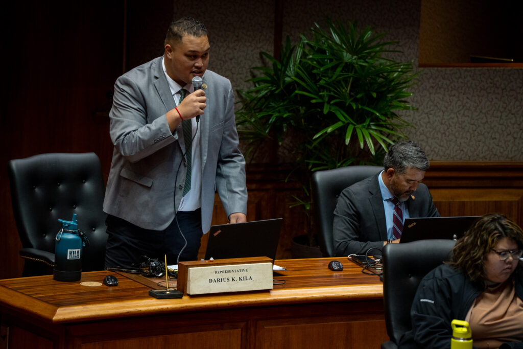 a man wearing a short gray blazer holds a microphone in his right hand as he speaks. He has short dark hair and is standing at a wooden desk with a name plate in front of him. Another man is seated with his head down at right, and a woman is seated at the lower right.