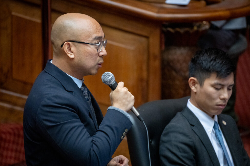 A balding man wearing glasses holds a microphone in his right hand as he looks right while speaking. Another man wearing a dark suit looks down.