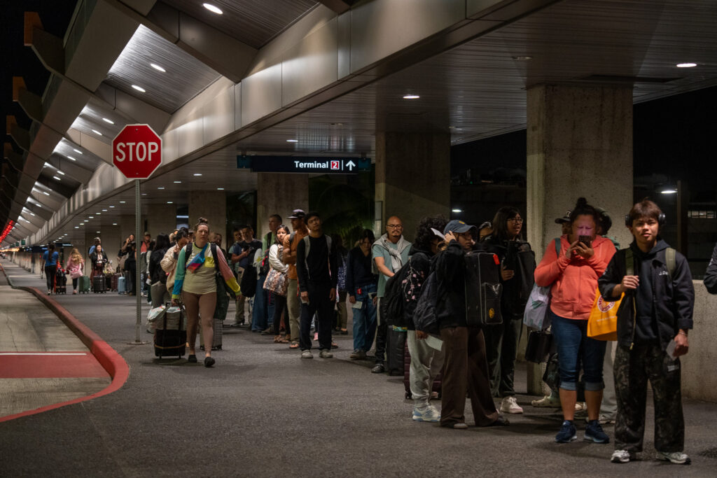 A ragged line of individuals waiting in a line next to concrete pillars to the right and a stop sign to the left. A woman wearing a light, multi-colored long sleeve top walks towards the camera with a roller bag pulled by her right hand.