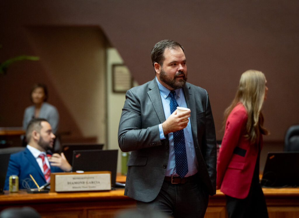 A man with short brown hair and beard walks through a room holding a paper cup. He is wearing a gray suit and blue tie. Another individual is seated at left behind him, and another individual with long brown hair and red blazer is in the background to the right.