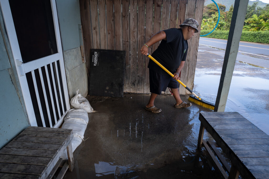 A man wearing dark clothes sweeps water on a sidewalk next to a white screen door with sandbags at its base.