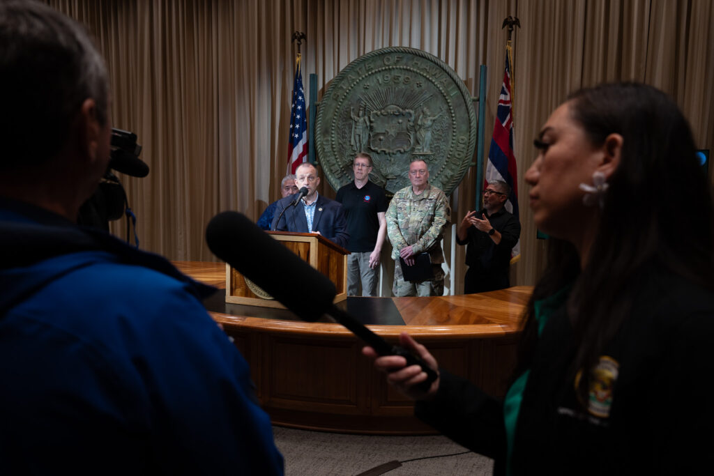 Several individuals stand near a lecturn with the Hawaii State Seal and flags behind them. They are framed by two individuals in the foreground.