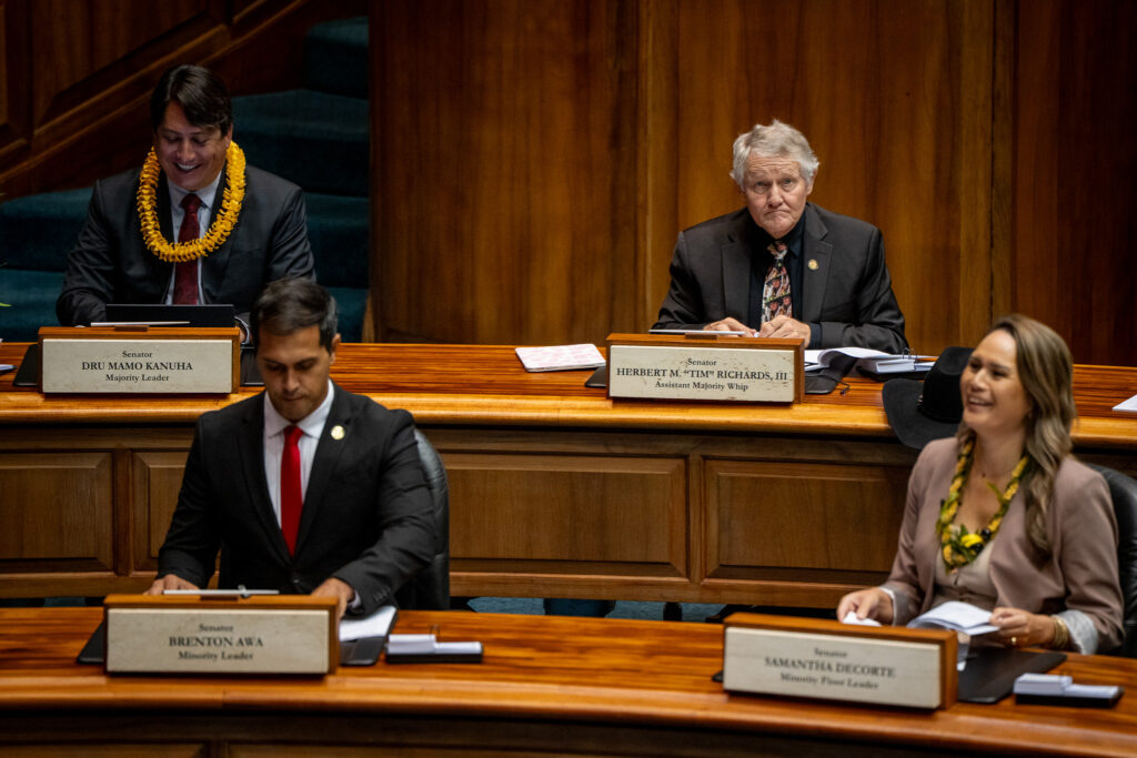 Four individuals sitting at parallel desks placed one in front of the other. The three men at left wear dark blazers and ties, and the woman at right wears a medium colored blazer.