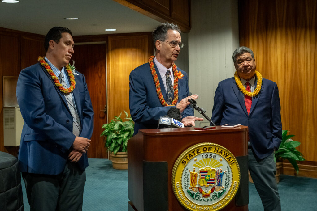 A man speaks at a lecturn as he is flanked by two other men. All wear dark blazers and lei.