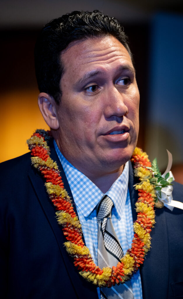 A head and shoulders photo of a man with short dark hair. He wears a checked blue shirt, diagonal gray tie, blue blazer and a yellow and orange lei.