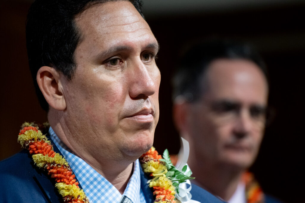 A horizontal photo showing two men from the neck up. The man in front is sharply focused while the man behind him is out of focus.