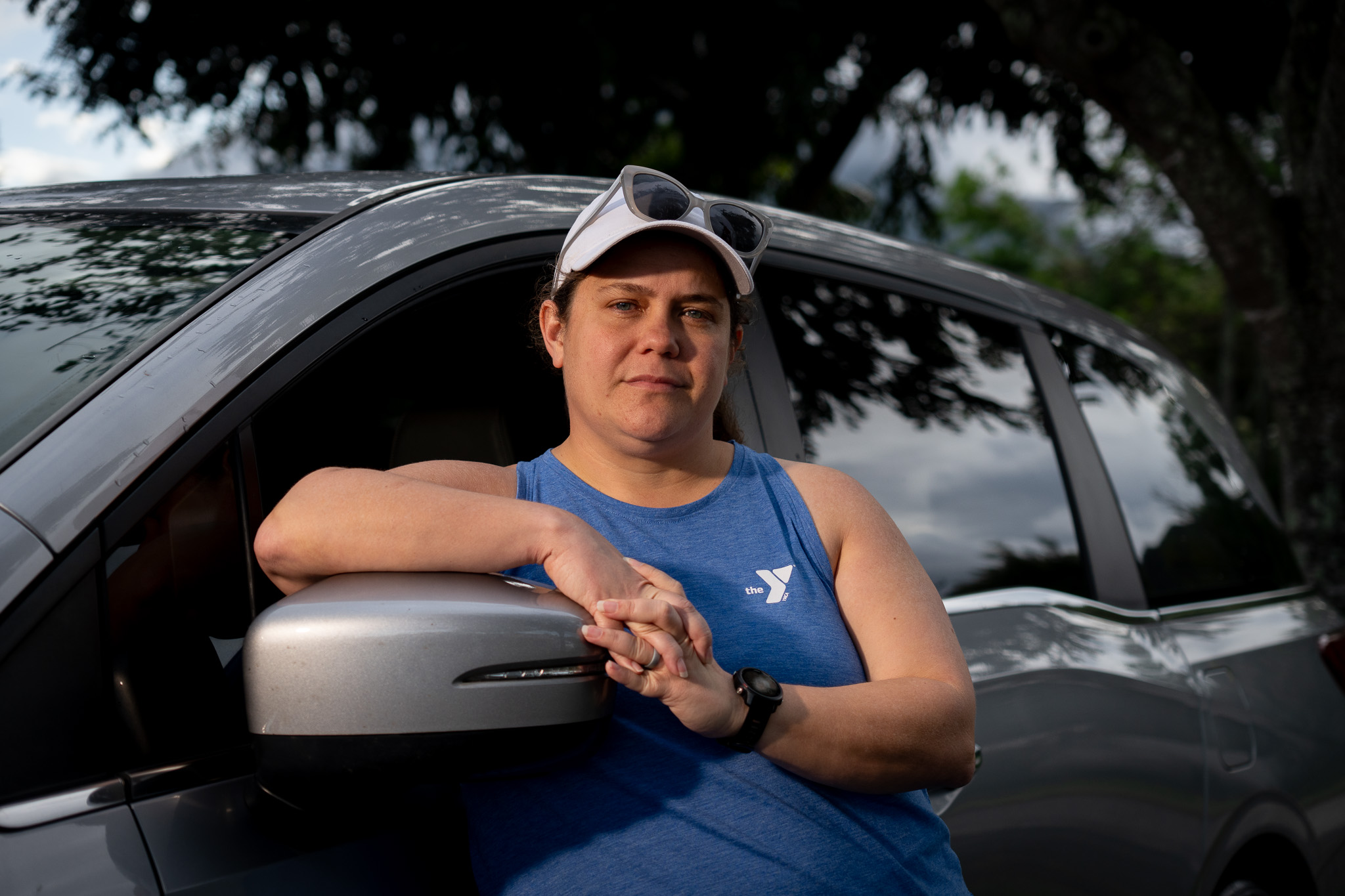 A woman is photographed wearing a white cap, sunglasses on her head and a blue tank top. She leans against a gray van.