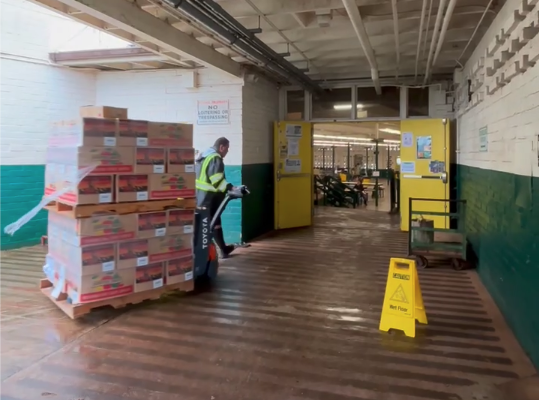 A man in neon jacket wheels a stack of supplies past a "wet floor" sign and into a large room at Leilehua High School in Wahiawā.