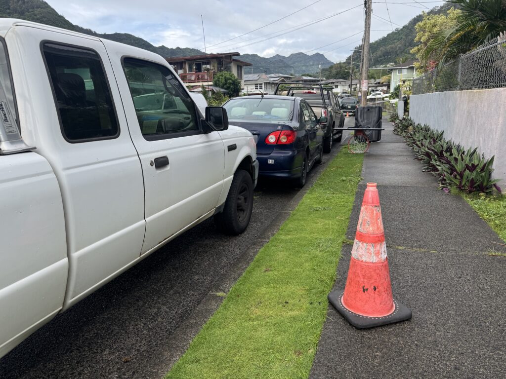 An orange cone sits on the sidewalk next to a car and a truck parked tightly in a Kalihi Valley neighborhood