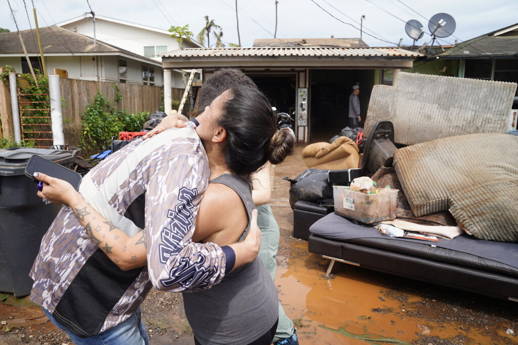 One Love Church volunteer Beau O’Brien of Honolulu hugs  Sharmaine Arial after helping move heavy, water damaged appliances and furniture out of her Kukea Loop home Sunday, March 22, 2026, in Waialua. Sharmaine’s number is 808-970-4295. (Kevin Fujii/Civil Beat/2026)
