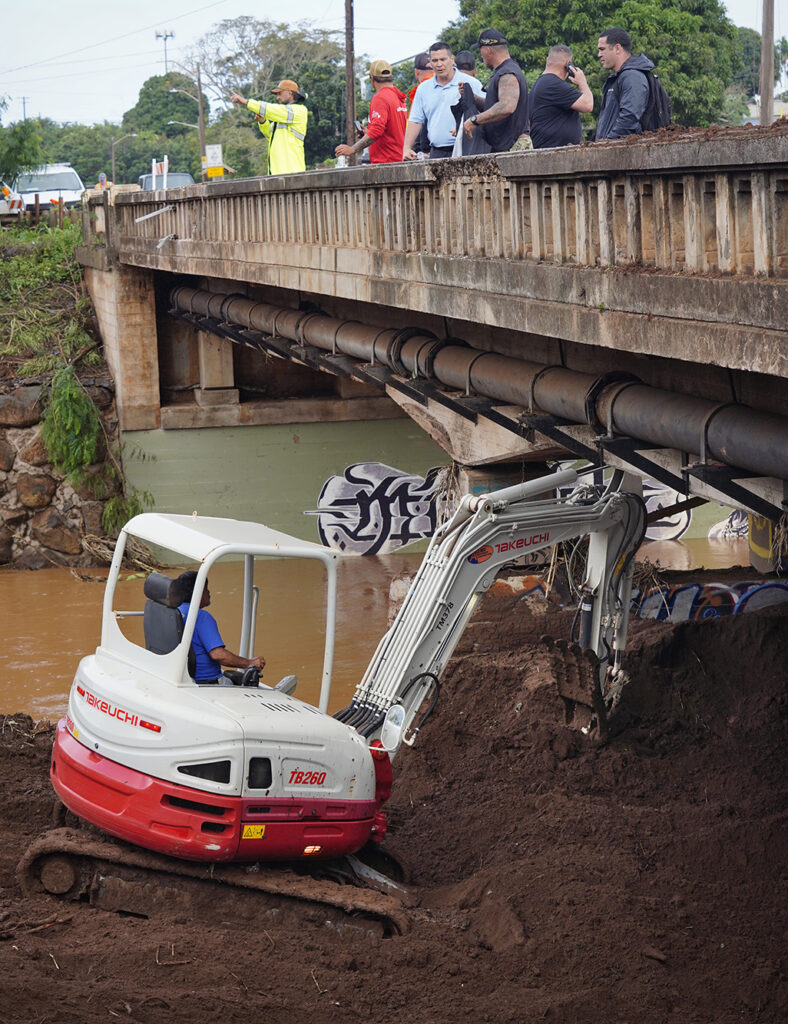 A privately owned compact excavator works to remove silt blocking a channel under the Otake Bridge as Department of Transportation director Ed Sniffen, blue shirt, talks story with community members and others over the Kaukonahua Stream Wednesday, March 25, 2026, in Waialua. (Kevin Fujii/Civil Beat/2026)