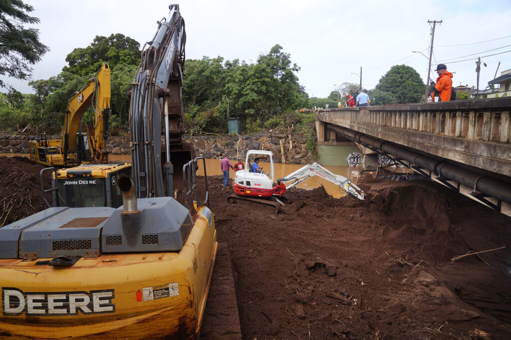 A compact excavator works to remove silt blocking a Kaukonahua Stream channel under the Otake Bridge as larger hydraulic crawler excavators wait for more room under the bridge to work Wednesday, March 25, 2026, in Waialua. All heavy machinery are privately owned. (Kevin Fujii/Civil Beat/2026)