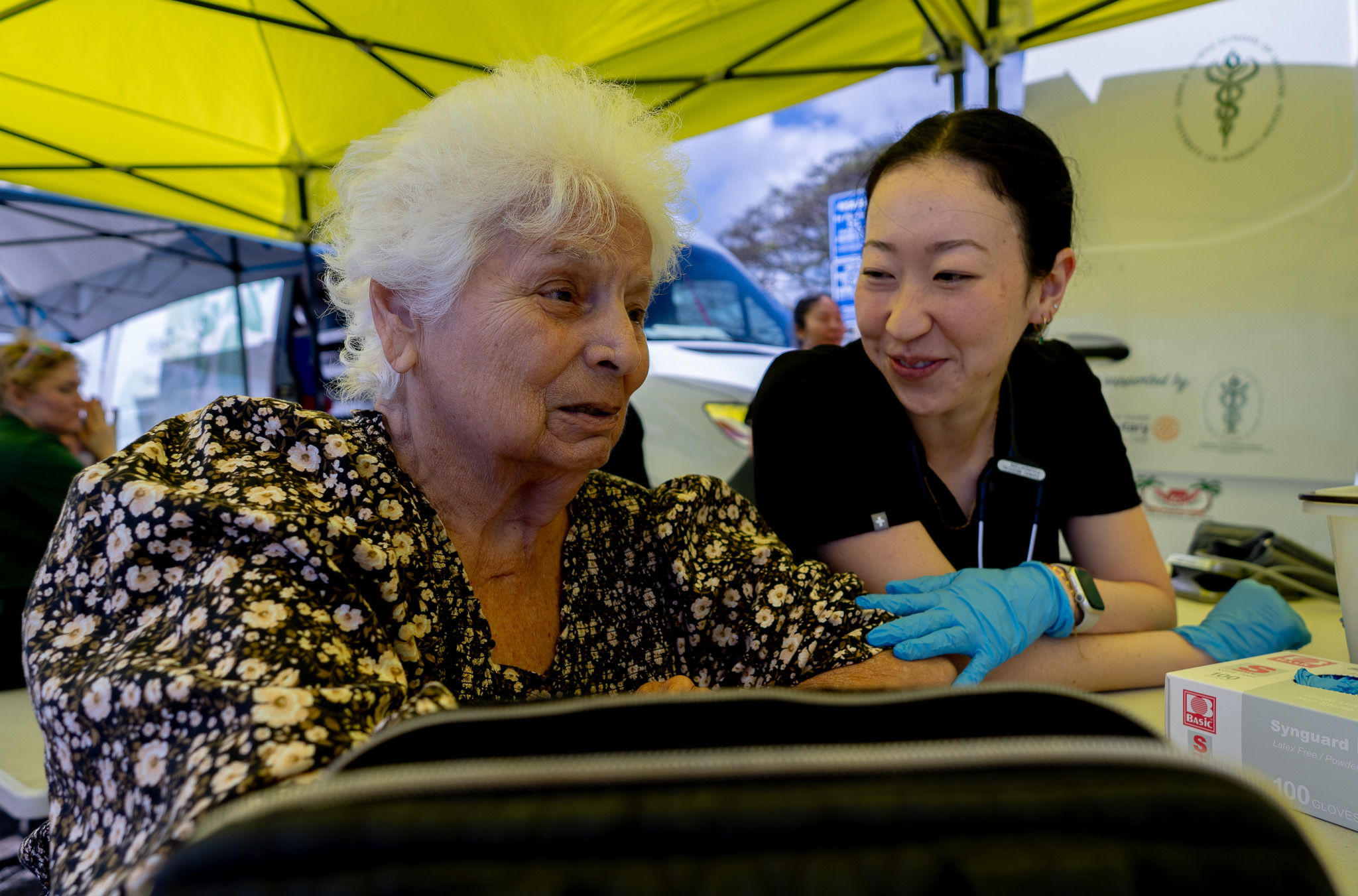 Haleiwa resident Sylvia Paulsen, 85, jokes with student doctor Kira Morikawa at a mobile clinic at Ali'i Beach Park March 26, 2026. Students from John A. Burns School of Medicine (JABSOM) staffed a clinic for area residents affected by flooding in Haleiwa and Waialua. (Craig Fujii/Civil Beat/2026)