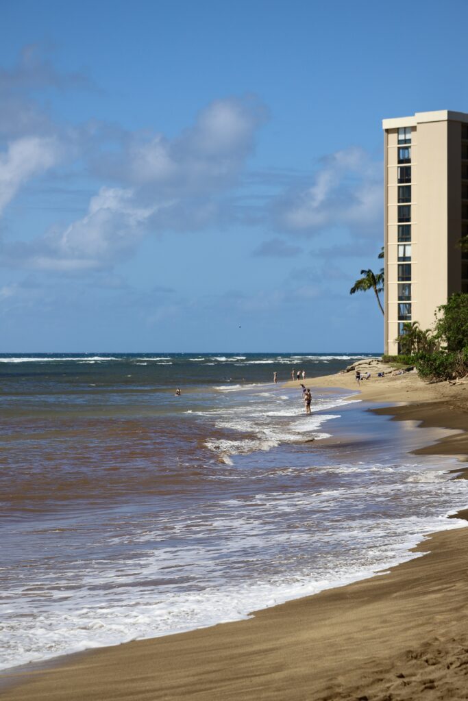 Several people in bathing suits swim and stand on a beach. There is a 12-story beige building on the beach. The ocean is blue, but there are large brown plumes.
