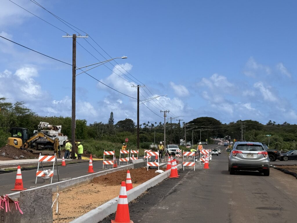 A car waits while orange cones surround a newly constructed cement median in the middle of the road. On the side, workers in bright yellow shirts look toward the ground.