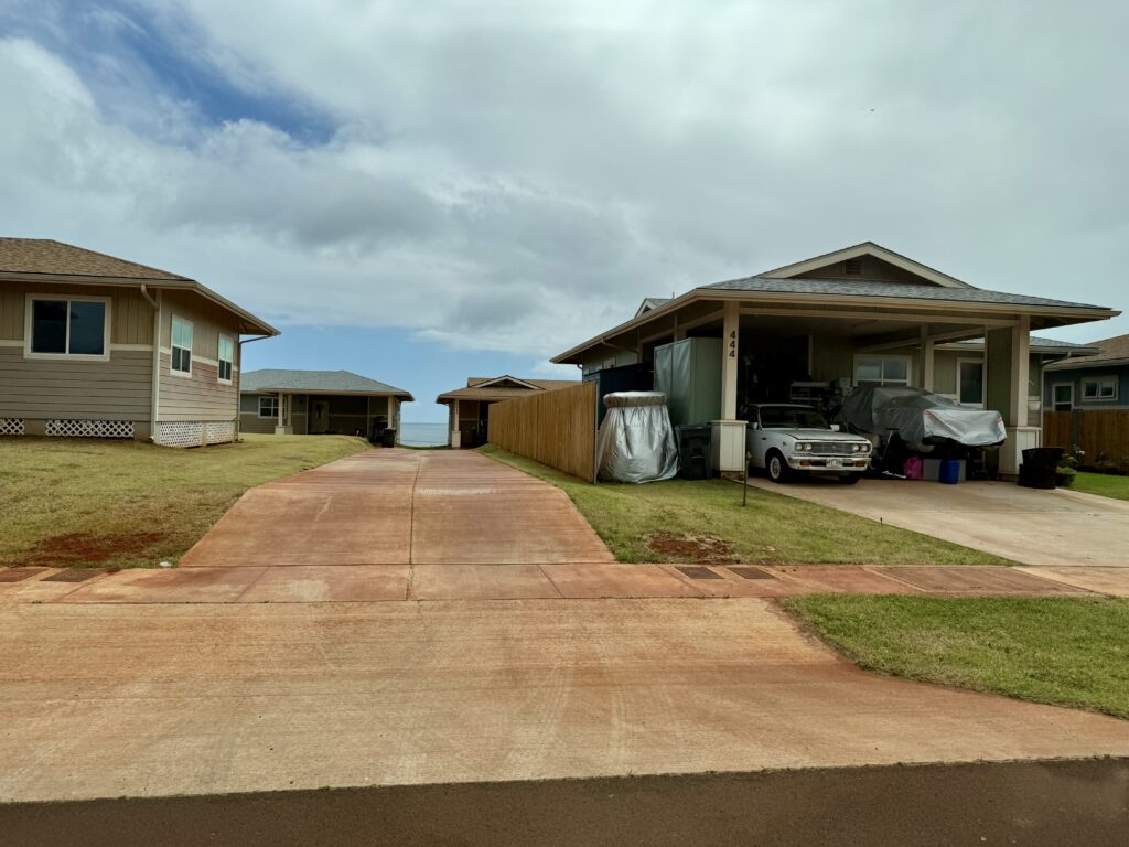 Two cars sit in an open carport. To the left, a driveway leads to two more houses in the background.