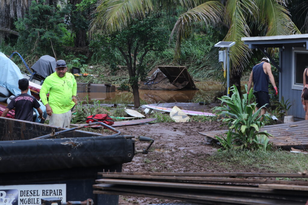 A man in a bright shirt standing next to a boy in the foreground with a dumpster bin and planks in the foreground. In the background is mud and a large muddy pool of water with garbage wheelie bins floating in it.
