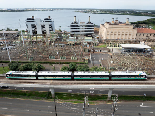 A four car train passes in front of several buildings which house an electricity generating station.