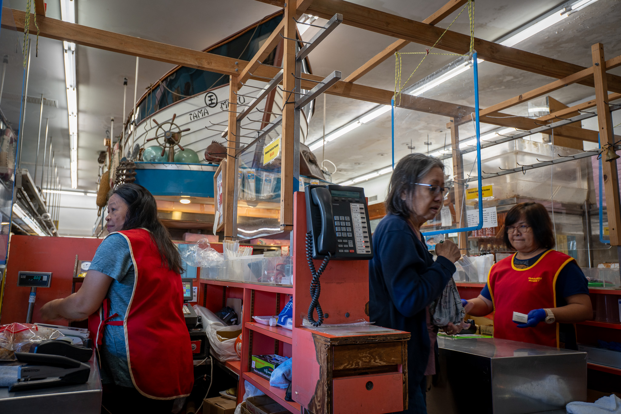 Jenny Ancheta, left, and Evelyn Favor staff the cash registers at Tamashiro Market April 3, 2026. Ancheta has worked at the store for almost 35 years, and Favor for just over a year. A large mockup of a boat dominates the floorspace, background.